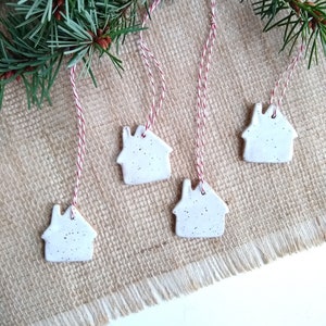 May include: White ceramic house ornaments with speckled detailing, hanging from red and white twine. The ornaments are displayed with a backdrop of a burlap surface and a sprig of evergreen branches, creating a festive holiday theme.