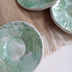 May include: Three handmade ceramic dishes with a light green glaze and a textured leaf design. The dishes have a white rim and a small, central indentation. They are arranged on a burlap surface.