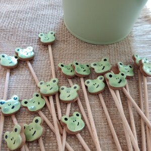 May include: A collection of green frog-shaped plant stakes with black facial details, arranged on a burlap surface. A light green bucket is in the background. The stakes are made of ceramic and wood.
