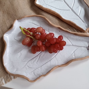 May include: Two white ceramic leaf-shaped serving platters with a speckled finish and a natural brown rim. One platter holds a bunch of red grapes. The platters are ideal for serving appetizers or displaying decorative items.