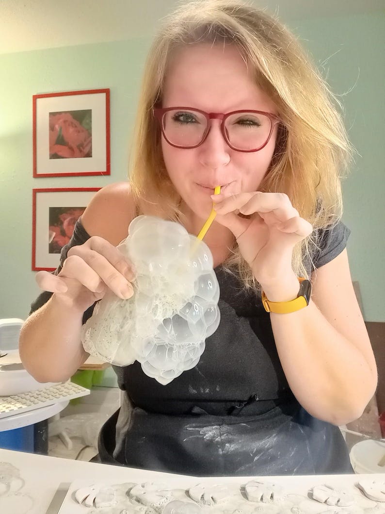 Peut inclure: Un moule en silicone translucide &agrave; la texture alv&eacute;ol&eacute;e en forme de grappe de raisin, tenu en main tandis qu'une paille jaune est utilis&eacute;e pour souffler de l'air &agrave; la surface afin de cr&eacute;er de la mousse. Le moule est souple et transparent.