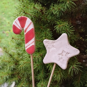 以下が含まれることがあります： 木製の棒に飾られた2つのセラミッククリスマスオーナメント。1つは赤と白のキャンディケーン、もう1つは雪の結晶模様の淡いピンクの星です。緑の松の葉を背景にしています。