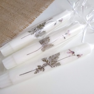 Op de afbeelding: Witte taps toelopende kaarsen versierd met geperste bloemen en bladeren. De kaarsen zijn verpakt in doorzichtig cellofaan en vastgebonden met witte linten. De bloemmotieven omvatten grijze bladeren en kleine roze bloemen.