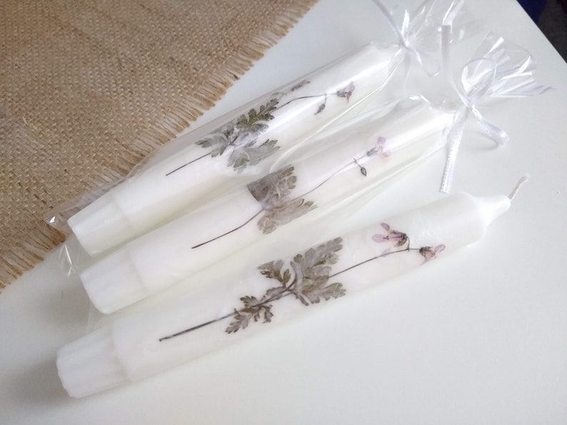 Op de afbeelding: Witte taps toelopende kaarsen versierd met geperste bloemen en bladeren. De kaarsen zijn verpakt in doorzichtig cellofaan en vastgebonden met witte linten. De bloemmotieven omvatten grijze bladeren en kleine roze bloemen.