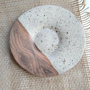May include: A handmade ceramic plate with a speckled white glaze and a brown, wood-grain textured design. The plate is round with a slightly raised center and a diameter of approximately 18 cm. The plate is a unique piece of pottery.