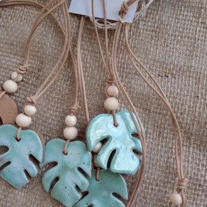Peut inclure: Une collection de colliers avec des pendentifs en c&eacute;ramique &eacute;maill&eacute;e en forme de feuille de monstera, arborant une finition bleu-vert brillante avec des taches sombres. Chaque pendentif est suspendu &agrave; un cordon beige avec deux perles en bois clair.