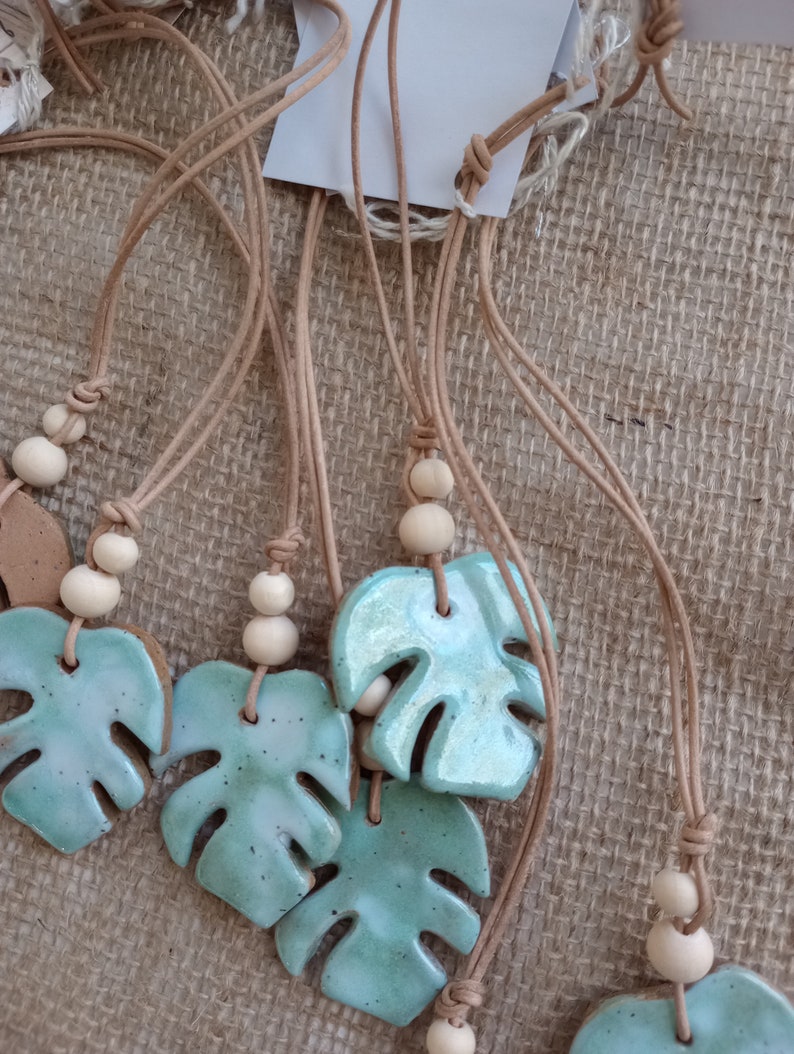 Peut inclure: Une collection de colliers avec des pendentifs en c&eacute;ramique &eacute;maill&eacute;e en forme de feuille de monstera, arborant une finition bleu-vert brillante avec des taches sombres. Chaque pendentif est suspendu &agrave; un cordon beige avec deux perles en bois clair.