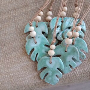 Peut inclure: Une collection de colliers avec des pendentifs en c&eacute;ramique en forme de feuilles de monstera. Chaque pendentif pr&eacute;sente un &eacute;mail vert menthe brillant et mouchet&eacute;, suspendu &agrave; un cordon beige orn&eacute; de deux perles en bois clair au-dessus.