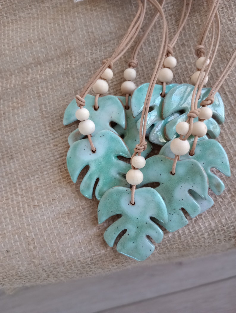 Peut inclure: Une collection de colliers avec des pendentifs en c&eacute;ramique en forme de feuilles de monstera. Chaque pendentif pr&eacute;sente un &eacute;mail vert menthe brillant et mouchet&eacute;, suspendu &agrave; un cordon beige orn&eacute; de deux perles en bois clair au-dessus.