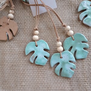 Peut inclure: Une collection de colliers avec des pendentifs en forme de feuille de monstera en c&eacute;ramique &eacute;maill&eacute;e dans des tons vert menthe et blanc. Chaque pendentif est suspendu &agrave; un cordon en cuir beige orn&eacute; de deux perles en bois clair.