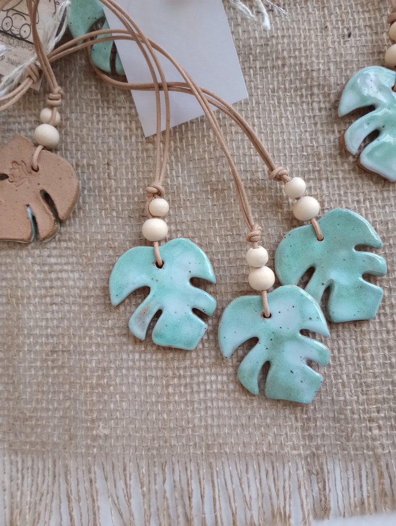 Peut inclure: Une collection de colliers avec des pendentifs en forme de feuille de monstera en c&eacute;ramique &eacute;maill&eacute;e dans des tons vert menthe et blanc. Chaque pendentif est suspendu &agrave; un cordon en cuir beige orn&eacute; de deux perles en bois clair.
