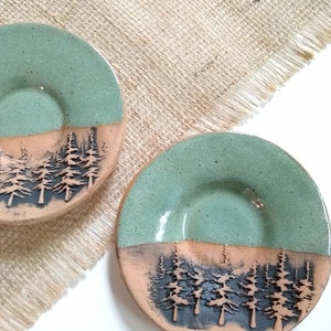 Op de afbeelding: Twee handgemaakte keramische schaaltjes. Elk schaaltje is rond met een lichtgroene bovenste helft en een bruinere onderste helft. De onderste helft toont een bos van donkere bomen. De schaaltjes liggen op een beige jute oppervlak.