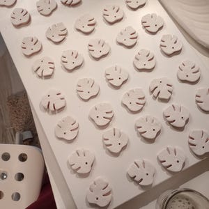 Peut inclure: Une collection de petits ornements en argile blanche en forme de feuille, dispos&eacute;s en grille sur une surface blanche. Chaque ornement pr&eacute;sente un motif d&eacute;coup&eacute; rappelant une feuille de monstera avec un trou pr&egrave;s du sommet pour la fixation.