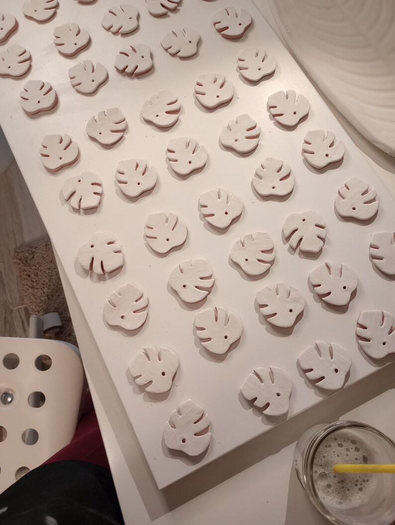 Peut inclure: Une collection de petits ornements en argile blanche en forme de feuille, dispos&eacute;s en grille sur une surface blanche. Chaque ornement pr&eacute;sente un motif d&eacute;coup&eacute; rappelant une feuille de monstera avec un trou pr&egrave;s du sommet pour la fixation.