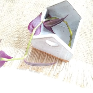 May include: A small, white ceramic house-shaped planter with a purple and green plant. The planter has a speckled texture and a rectangular opening. The plant's leaves are a deep purple color, and the stems are green.