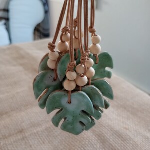 Peut inclure: Un ensemble de pendentifs en c&eacute;ramique en forme de feuille de monstera avec un &eacute;mail vert sauge mouchet&eacute;. Chaque feuille est suspendue par un cordon en cuir beige orn&eacute; de deux perles en bois clair, fix&eacute;es par des n&oelig;uds au-dessus de la c&eacute;ramique.