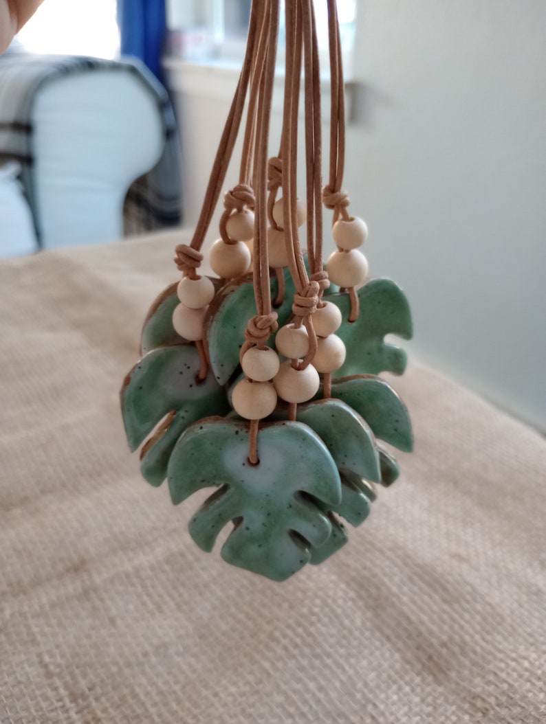 Peut inclure: Un ensemble de pendentifs en c&eacute;ramique en forme de feuille de monstera avec un &eacute;mail vert sauge mouchet&eacute;. Chaque feuille est suspendue par un cordon en cuir beige orn&eacute; de deux perles en bois clair, fix&eacute;es par des n&oelig;uds au-dessus de la c&eacute;ramique.