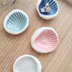 May include: Four small, ceramic seashell-shaped dishes in shades of blue, pink, and white. One dish holds a silver ring. The dishes have a textured, ridged interior and are displayed on a wooden surface. These are ideal for holding jewelry.