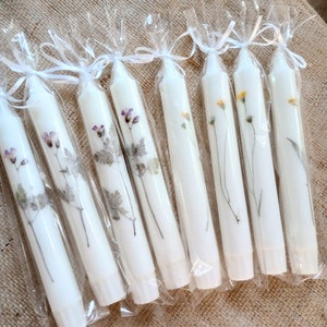 Op de afbeelding: Witte taps toelopende kaarsen versierd met geperste paarse en gele wilde bloemen. Elke kaars is verpakt in doorzichtig cellofaan en vastgebonden met een wit lint. De tekst "Purple wildflower" en "Yellow wildflower" is zichtbaar.