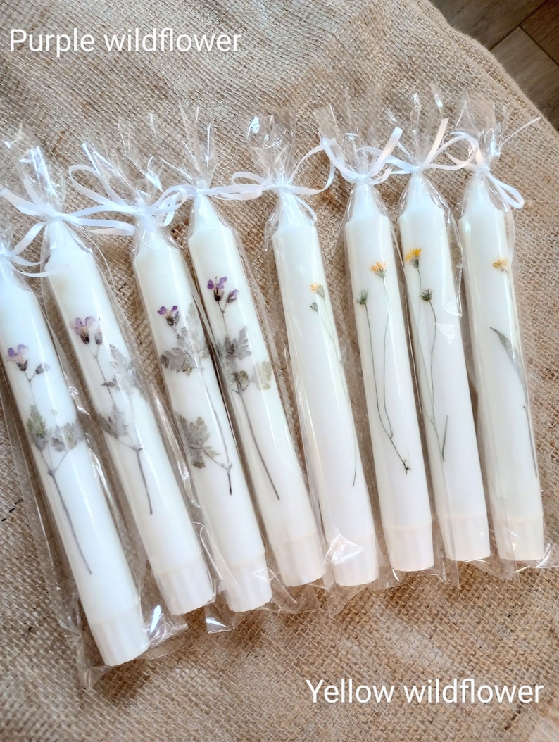 Op de afbeelding: Witte taps toelopende kaarsen versierd met geperste paarse en gele wilde bloemen. Elke kaars is verpakt in doorzichtig cellofaan en vastgebonden met een wit lint. De tekst "Purple wildflower" en "Yellow wildflower" is zichtbaar.