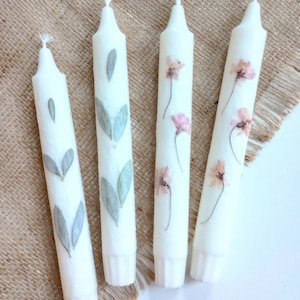 Op de afbeelding: Vier witte taps toelopende kaarsen. Twee kaarsen hebben olijfbladmotieven en twee hebben kersenbloesemmotieven. De kaarsen zijn versierd met geperste bladeren en bloemen. De tekst "Olive leaf" en "Cherry blossom" is zichtbaar.