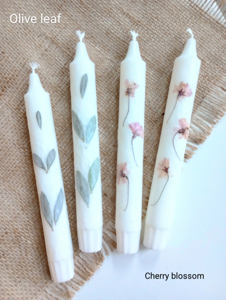 Op de afbeelding: Vier witte taps toelopende kaarsen. Twee kaarsen hebben olijfbladmotieven en twee hebben kersenbloesemmotieven. De kaarsen zijn versierd met geperste bladeren en bloemen. De tekst "Olive leaf" en "Cherry blossom" is zichtbaar.