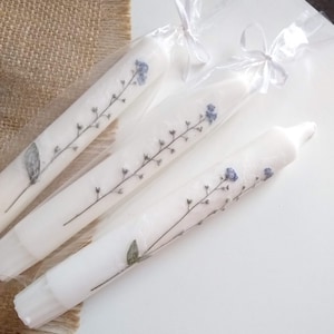 Op de afbeelding: Drie witte dinerkaarsen versierd met delicate blauwe en groene bloemtakken. Elke kaars is verpakt in transparant cellofaan en vastgebonden met een wit lint. De kaarsen worden gepresenteerd op een wit oppervlak, met een jute achtergrond.