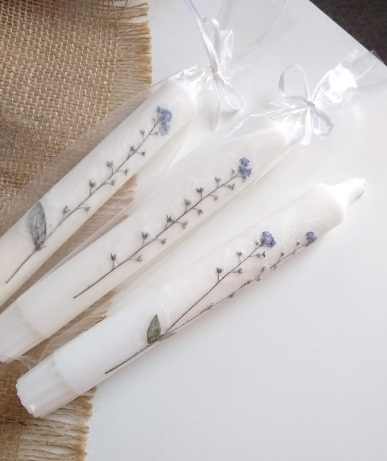 Op de afbeelding: Drie witte dinerkaarsen versierd met delicate blauwe en groene bloemtakken. Elke kaars is verpakt in transparant cellofaan en vastgebonden met een wit lint. De kaarsen worden gepresenteerd op een wit oppervlak, met een jute achtergrond.