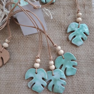 Peut inclure: Plusieurs pendentifs en c&eacute;ramique en forme de feuilles de monstera avec un &eacute;mail vert menthe brillant et des taches sombres. Chaque pendentif est suspendu &agrave; un cordon en cuir beige orn&eacute; de deux petites perles en bois clair au-dessus du motif.
