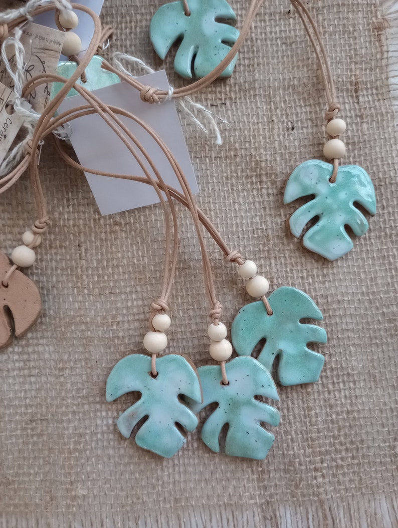Peut inclure: Plusieurs pendentifs en c&eacute;ramique en forme de feuilles de monstera avec un &eacute;mail vert menthe brillant et des taches sombres. Chaque pendentif est suspendu &agrave; un cordon en cuir beige orn&eacute; de deux petites perles en bois clair au-dessus du motif.