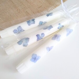 Op de afbeelding: Vier witte stompkaarsen versierd met geperste blauwe hortensiabloemen, verpakt in transparant cellofaan en vastgebonden met een wit lint. De kaarsen zijn een decoratief item.