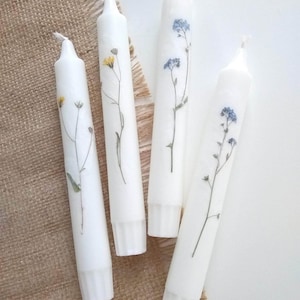 Op de afbeelding: Vier witte dinerkaarsen versierd met geperste bloemen. Twee kaarsen hebben blauwe vergeet-mij-nietjes en twee gele wilde bloemen. De kaarsen zijn gerangschikt op een gestructureerd, neutraal gekleurd oppervlak.