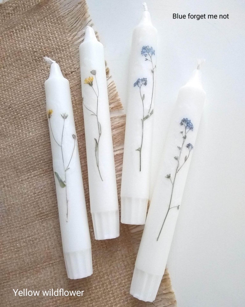 Op de afbeelding: Vier witte dinerkaarsen versierd met geperste bloemen. Twee kaarsen hebben blauwe vergeet-mij-nietjes en twee gele wilde bloemen. De kaarsen zijn gerangschikt op een gestructureerd, neutraal gekleurd oppervlak.