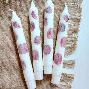 Op de afbeelding: Vier witte taps toelopende kaarsen versierd met bordeauxrode rozenblaadjes. De kaarsen zijn gerangschikt op een jute oppervlak. De tekst "Burgundy Rose" staat onderaan de afbeelding.