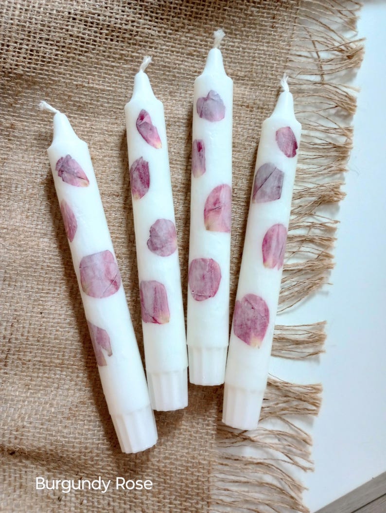 Op de afbeelding: Vier witte taps toelopende kaarsen versierd met bordeauxrode rozenblaadjes. De kaarsen zijn gerangschikt op een jute oppervlak. De tekst "Burgundy Rose" staat onderaan de afbeelding.