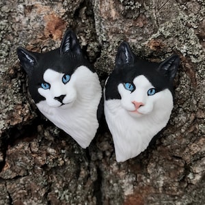 May include: Two black and white cat head sculptures with blue eyes. The sculptures are made of a white material and are sitting on a piece of tree bark.