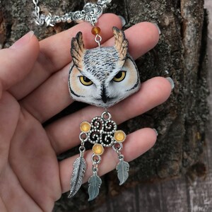 Puede incluir: Un collar de cadena plateada con un colgante de b&uacute;ho detallado hecho de arcilla polim&eacute;rica. El b&uacute;ho tiene plumas realistas marrones, blancas y negras y ojos amarillos. El colgante est&aacute; unido a un amuleto atrapasue&ntilde;os plateado con cuentas amarillas y plumas.