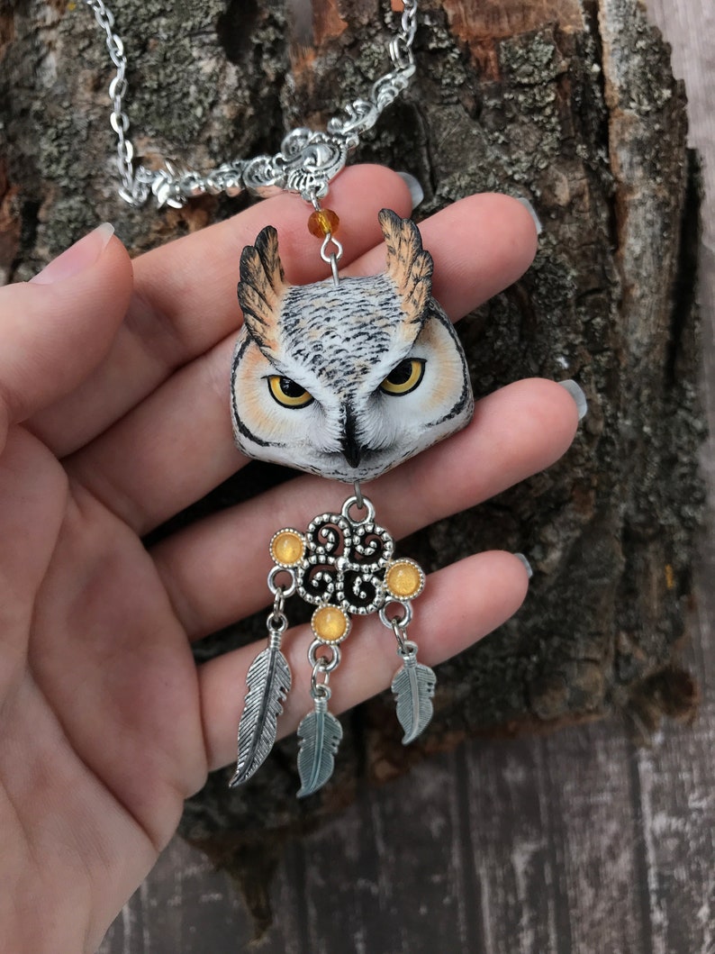 Puede incluir: Un collar de cadena plateada con un colgante de b&uacute;ho detallado hecho de arcilla polim&eacute;rica. El b&uacute;ho tiene plumas realistas marrones, blancas y negras y ojos amarillos. El colgante est&aacute; unido a un amuleto atrapasue&ntilde;os plateado con cuentas amarillas y plumas.