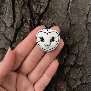 May include: A white ceramic owl head with black and yellow details. The owl has black eyes with yellow pupils and a black beak. The owl is sitting on a hand.