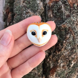May include: A small ceramic owl figurine with brown and white markings. The owl has black eyes and a white beak.