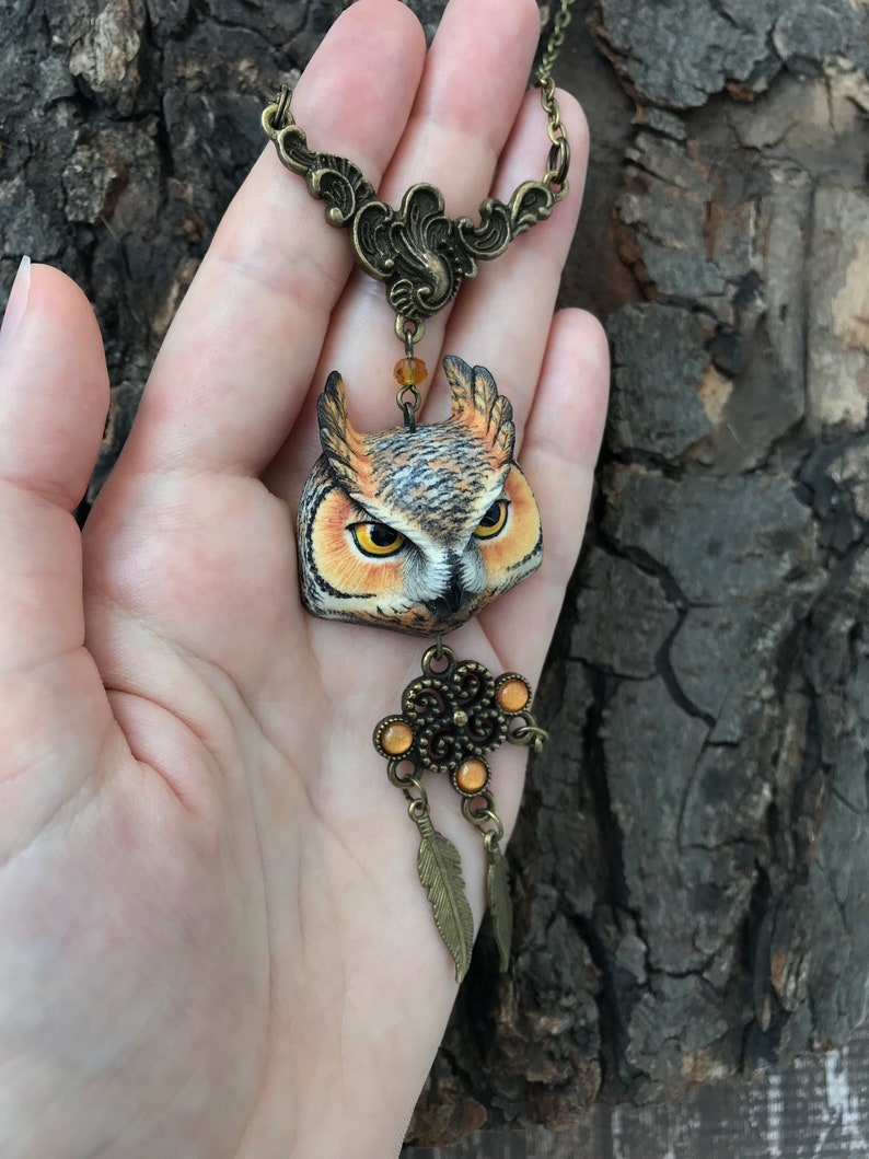 Puede incluir: Un collar con un colgante de b&uacute;ho detallado, pintado en marr&oacute;n, blanco y naranja. El colgante est&aacute; unido a una cadena con una filigrana de bronce decorativa y un peque&ntilde;o amuleto con piedras de color &aacute;mbar.