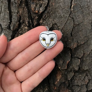 May include: A small white ceramic owl figurine with black and yellow details. The owl has black eyes and a yellow beak.