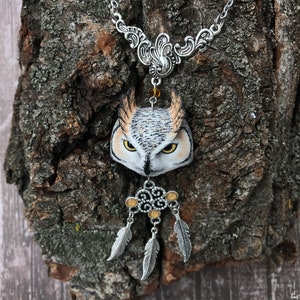 Puede incluir: Un collar de cadena de plata con un colgante de b&uacute;ho detallado y un amuleto de atrapasue&ntilde;os de plata con tres plumas. El b&uacute;ho est&aacute; pintado con detalles realistas, incluyendo plumas marrones, blancas y amarillas.