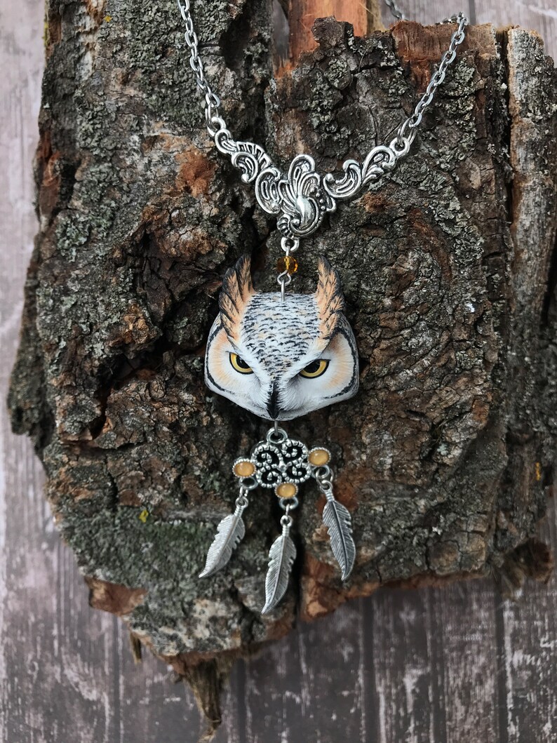 Puede incluir: Un collar de cadena de plata con un colgante de b&uacute;ho detallado y un amuleto de atrapasue&ntilde;os de plata con tres plumas. El b&uacute;ho est&aacute; pintado con detalles realistas, incluyendo plumas marrones, blancas y amarillas.