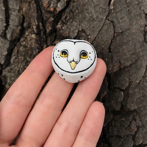 May include: A white ceramic owl figurine with black eyes and a yellow beak. The owl is painted with black dots and has a white background.
