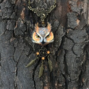 Puede incluir: Un collar con un colgante de b&uacute;ho marr&oacute;n con un dise&ntilde;o de atrapasue&ntilde;os. El b&uacute;ho tiene ojos naranjas y est&aacute; hecho de arcilla polim&eacute;rica. El collar est&aacute; hecho de cadena de bronce y tiene un amuleto de atrapasue&ntilde;os de bronce con cuentas naranjas.