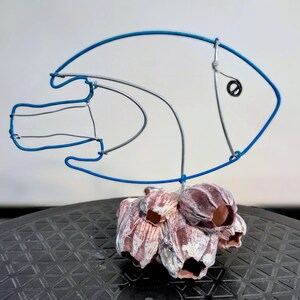 May include: A wire sculpture of a fish with a blue outline and a black eye. The fish is mounted on a cluster of barnacles with pink and white hues. The sculpture is a decorative item.