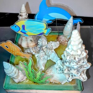 May include: A decorative ocean-themed display featuring a blue dolphin and colorful fish figurines. The display includes seashells, green seaweed, and a sandy base, all encased in a rectangular frame. The fish are yellow and orange.