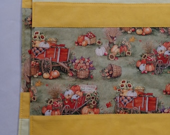 May include: Two green fabric placemats with a fall harvest theme. The placemats feature a yellow, green, and white striped pattern with a repeated image of a red wagon filled with pumpkins, gourds, and apples. The text "Pumpkins, Apple Cider, Hayrides" is printed on a sign in the image.