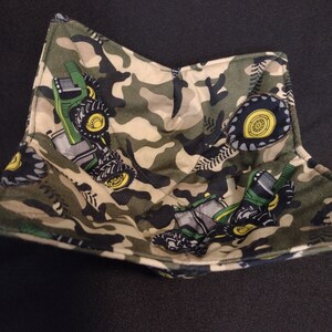 May include: A fabric bowl with a camouflage pattern featuring green and yellow tractors. The bowl is square-shaped with a quilted design. The background is a solid black.